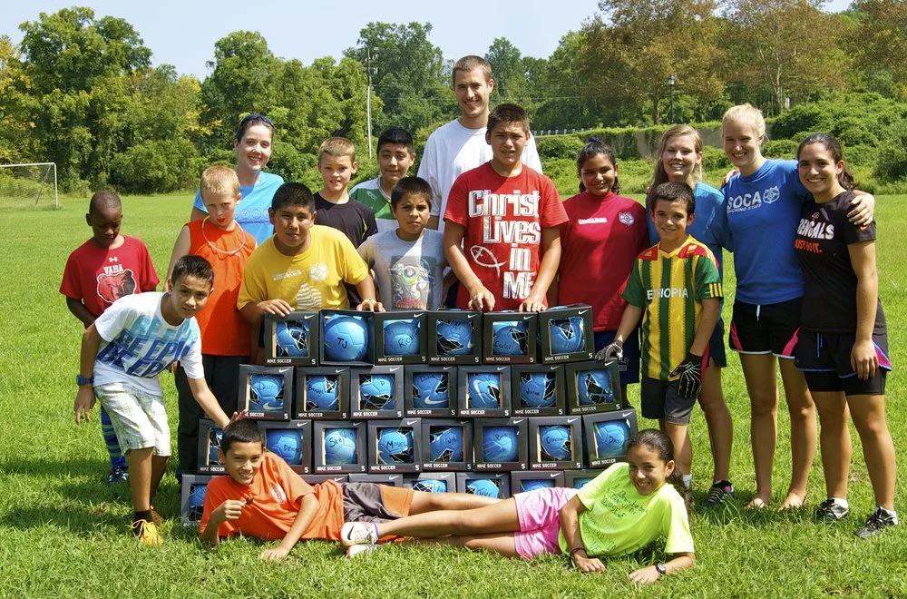 Kids and leaders from the Southwood soccer camp posing behind a stack of new soccer balls on the field
