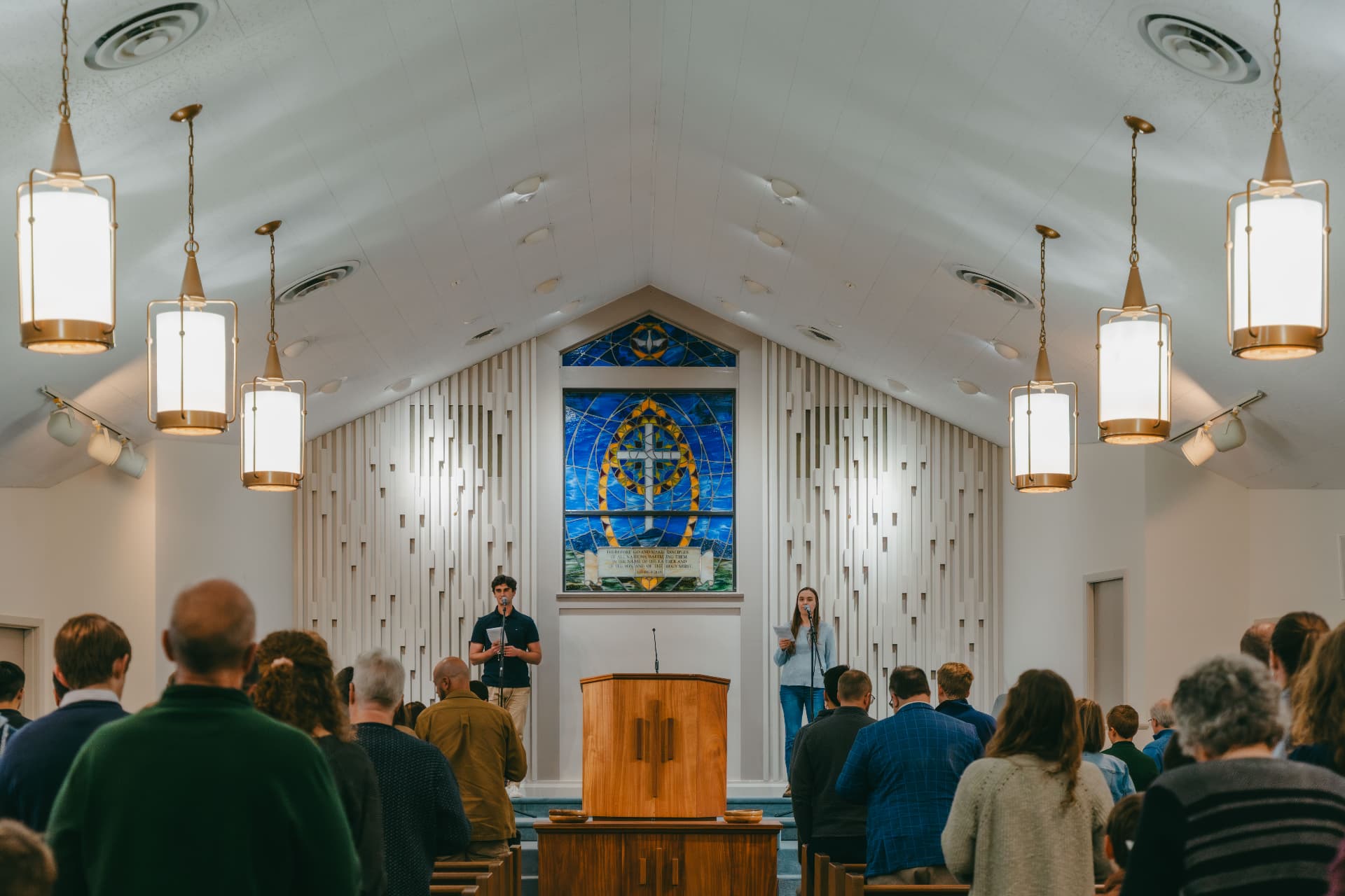 The congregation standing in worship in the sanctuary, facing the wooden pulpit and the stained glass cross window